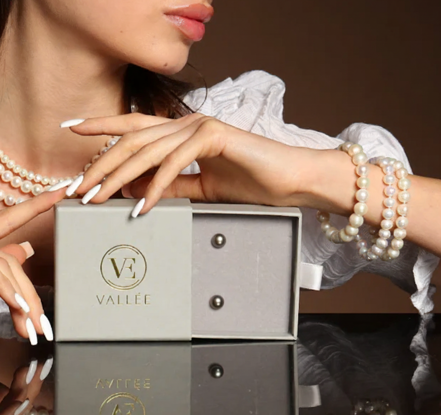 Frau präsentiert eine geöffnete Vallée Schmuckbox mit Tahitit-Perlenohrringen, trägt Perlenarmbänder und eine Perlenkette.